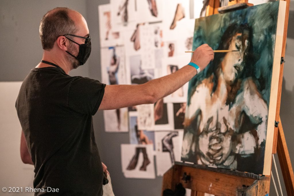 Artist Dave Clay standing in front of a canvas painting a woman posed provocatively, reference images visible on wall behind him.