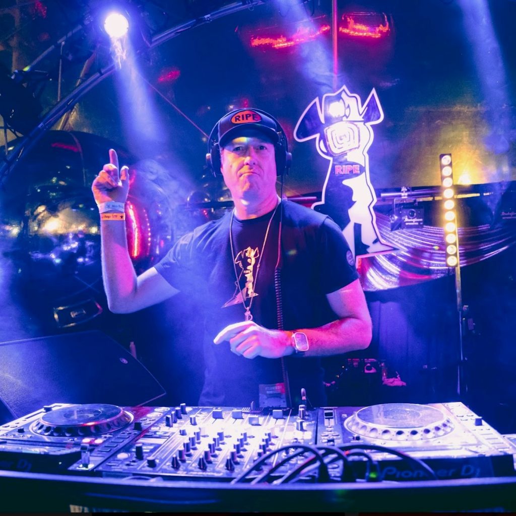 Color picture of Deckard standing in front of a mixer, hands up in classic iconography pose, wearing a baseball hat and t-shirt.