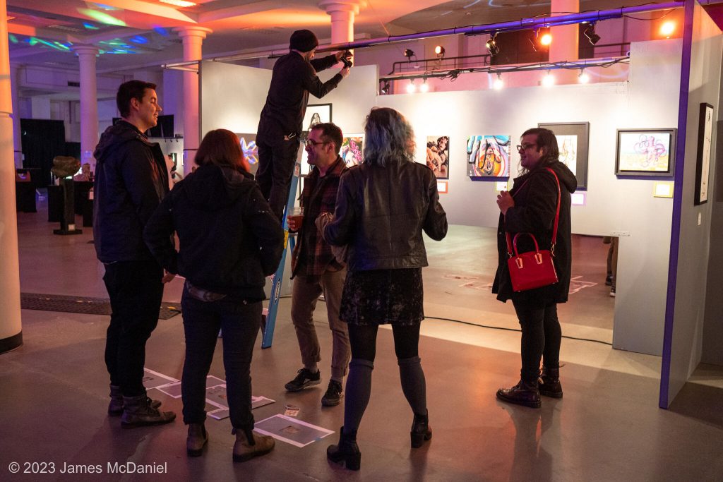 Five people standing around chatting, next to a person on a ladder adjusting lights over art work.