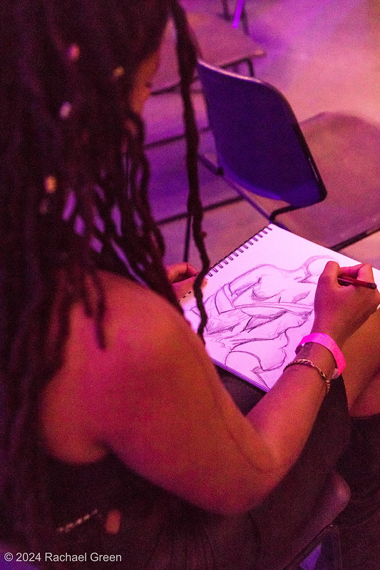 2025 Call for Art: A photo over the shoulder of a black woman with long hair who is drawing a pencil sketch of a woman, taken during one of the Life Drawing sessions of SEAF 2024.